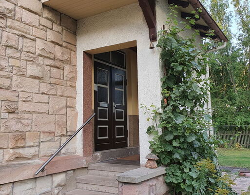 Der überdachte Eingang zum Pfarrbüro. Vor der Tür geht es fünf Stufen hinauf. Links daneben an der Hauswand ein Geländer.